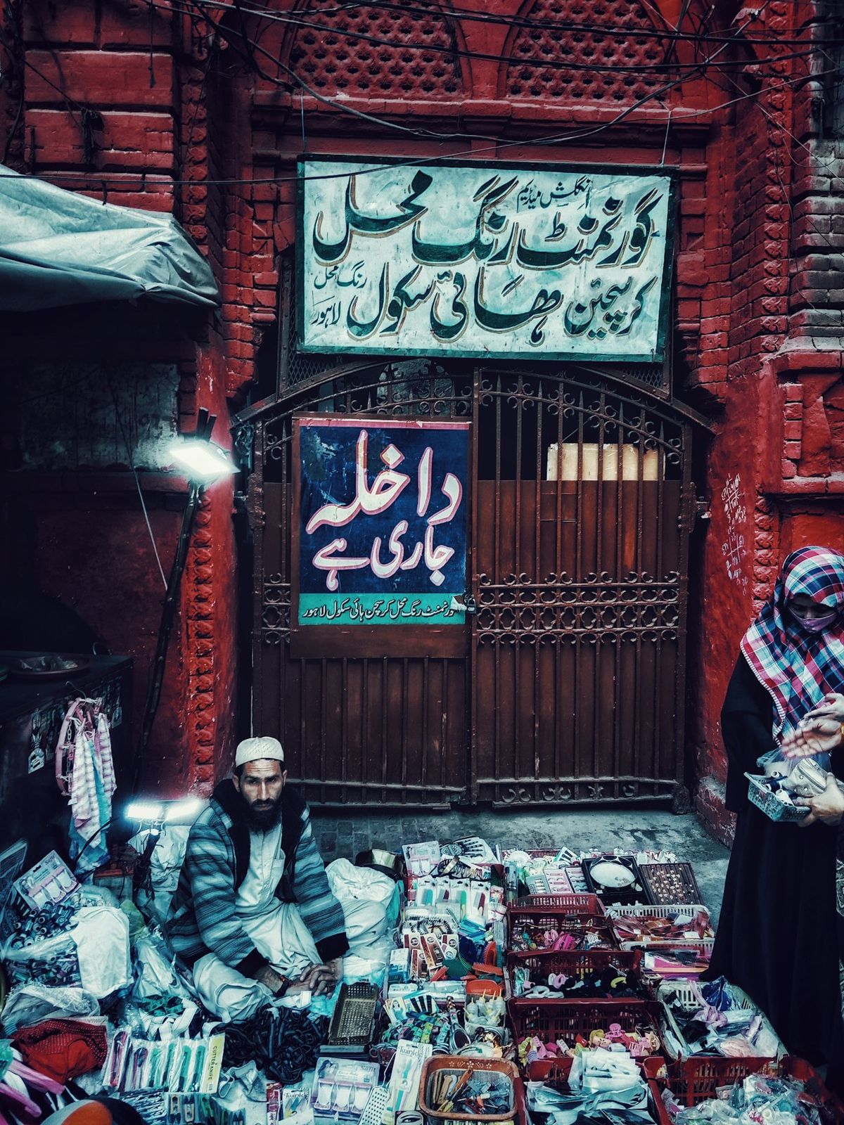 A shopkeeper sitting among his goods in front of a shop with Urdu signage